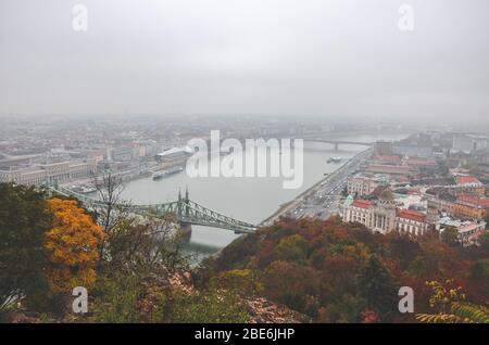 Budapest, Ungarn - 6. November 2019: Ungarische Hauptstadt Stadtbild mit Donau und Freiheitsbrücke. Die Altstadt im Hintergrund. Bewölktes regnerisches Wetter. Bunte Herbstbäume im Vordergrund. Stockfoto