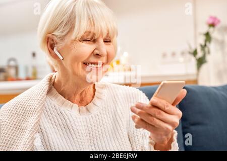 Graue Haare Frau mit Ohrhörern mit modernen Smartphone, verbringen Tag zu Hause Stockfoto