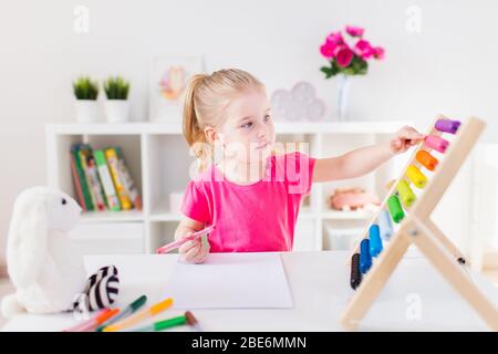 Kleines lächelndes blondes Mädchen, das am weißen Schreibtisch sitzt und auf den bunten Abakus im Klassenzimmer zählt. Bildung zu Hause. Stockfoto