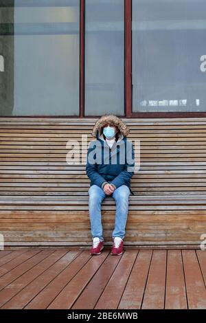 Kranker Mann mit Kapuze, der allein auf der Bank sitzt und eine schützende Gesichtsmaske gegen übertragbare Infektionskrankheiten trägt, covid-19. Leere Stadt von peopl Stockfoto