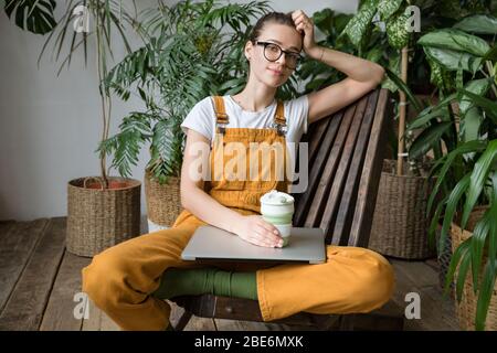 Europäische Gärtnerin trägt Overalls, Ruhe nach der Arbeit, sitzt auf Holzstuhl im Haus Gewächshaus, halten wiederverwendbare Kaffee / Tee Tasse, Blick auf ca. Stockfoto