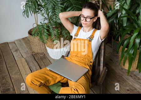 Gärtnerin legte Hände hinter den Kopf fühlt sich ruhig, Ruhe nach der Arbeit, sitzen auf Holzstuhl im Garten zu Hause, geschlossene Augen, Laptop auf den Knien.Br Stockfoto