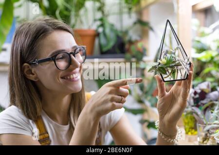 Lächelnde Frau Gärtner in Brille zeigt Zeigefinger auf Stecklinge von tradescantia in einem kleinen Glasflorarium für die Pflanzenkeimung, eingemachtes Houseplan Stockfoto