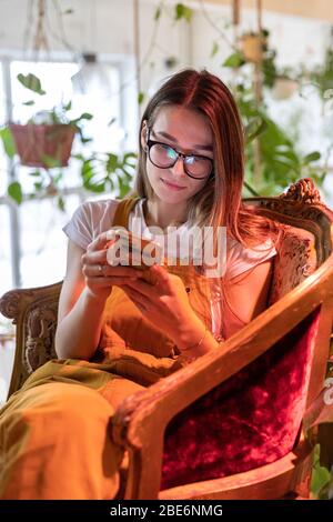 Junge Gärtnerin in Gläsern trägt Overalls, sitzt auf Vintage-Stuhl im Gewächshaus und mit Smartphone, Eingabe Nachricht, selektive Fokussierung. Werk Stockfoto