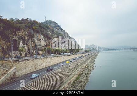 Budapest, Ungarn - 6. November 2019: Gellert Hill Cave Church, Sziklatemplom. Freiheitsstatue auf dem Hügel. Autos auf der Straße vor dem Hügel an der Donau. Ungarische Sehenswürdigkeiten. Stockfoto