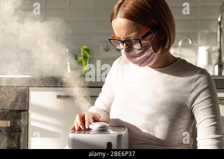 Frau im Gesicht Schutzmaske hält in ihren Händen arbeiten Aroma Öl Luftbefeuchter im sonnigen Raum. Befeuchtung der Luft in der Wohnung während der Perio Stockfoto