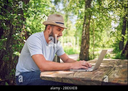 Bärtiger Mann arbeitet und lächelt mit Laptop im Park unter dem Baum. Happy Freelancer sitzt und nutzt App oder Website auf dem Rasen. Arbeit Stockfoto