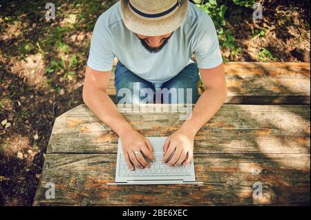 Bärtiger Mann arbeitet und lächelt mit Laptop im Park unter dem Baum. Happy Freelancer sitzt und nutzt App oder Website auf dem Rasen. Arbeit Stockfoto