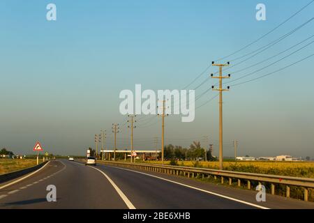 Perspektivische Aufnahme von Beton-Strompolen und Drähten in der Nähe der Autobahn auf dem ländlichen Raum. Das Morgenlicht sorgt für eine warme Atmosphäre. Stockfoto