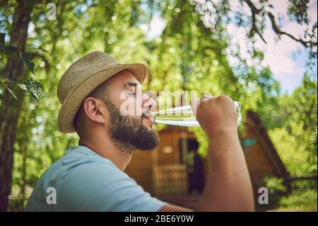 Porträt eines glücklichen jungen Mannes, der während des Sitzens und Ausruhens in einem Park Wasser aus einer Flasche trinkt Stockfoto