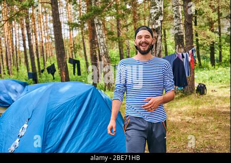 Fröhlicher junger, bärtiger Mann, der draußen vor dem Hintergrund eines Touristenzeltes campt. Stockfoto