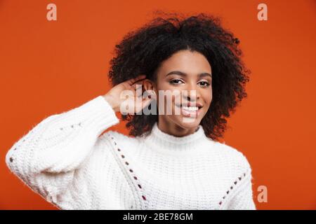 Schöne fröhliche junge afrikanerin trägt Pullover, die über rotem Hintergrund steht und versucht, Gerüchte zu hören Stockfoto