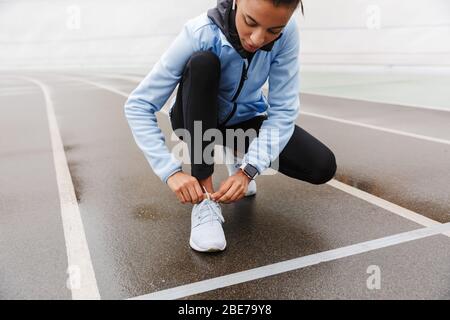 Zugeschnittenes Bild einer attraktiven jungen afrikanischen Sportlerin, die sich nach dem Laufen im Stadion ausruht und Schuhwerk bindet Stockfoto