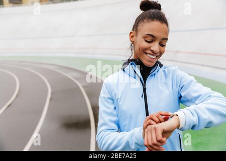 Attraktive junge afrikanische Sportlerin, die sich nach dem Rennen im Stadion ausruht und die Smartwatch anschaut Stockfoto