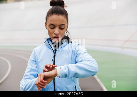 Attraktive junge afrikanische Sportlerin, die sich nach dem Rennen im Stadion ausruht und die Smartwatch anschaut Stockfoto