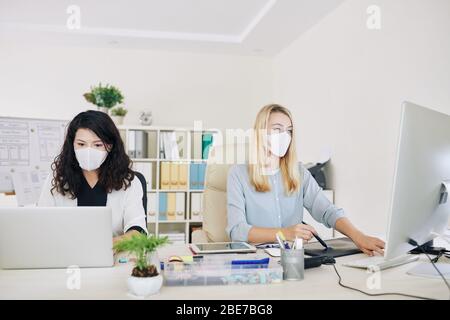 Team von Grafikerinnen in medizinischen Masken, die nebeneinander am Büroschreibtisch sitzen, wenn sie während der Pandemie an einem Projekt arbeiten Stockfoto