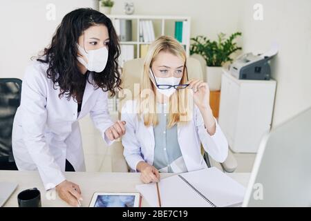 Junge medizinische Arbeiterinnen in Labormänteln und medizinischen Masken, die Röntgenaufnahmen der Brust auf dem Computerbildschirm analysieren Stockfoto