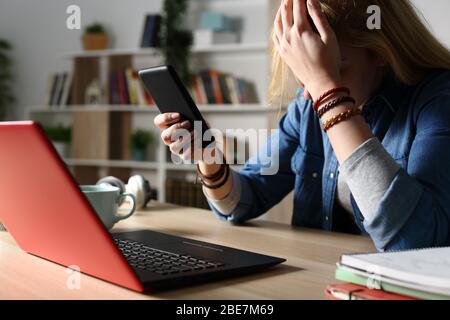 Nahaufnahme eines traurigen Studenten, der schlechte Nachrichten auf einem Smartphone liest, das nachts auf einem Schreibtisch sitzt Stockfoto