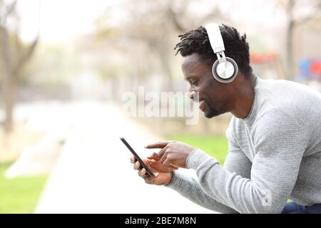 Seitenansicht Porträt eines schwarzen Mannes, der Musik auf Kopfhörern und Telefon auf einer Parkbank hört Stockfoto