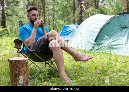 Junger Tourist Mann hält Tasse Kaffee auf Hintergrund des Zeltes am Morgen, genießen Ruhe und Freiheit im Wald. Stockfoto