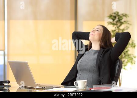 Entspannte leitende Frau, die frische Luft mit den Armen auf dem Kopf auf einem Schreibtisch im Büro atmet Stockfoto