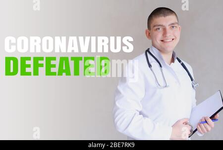 Junge kaukasische Arzt jubelt wegen der besiegten Coronavirus und stehend in grauen Wand Hintergrund. Stockfoto