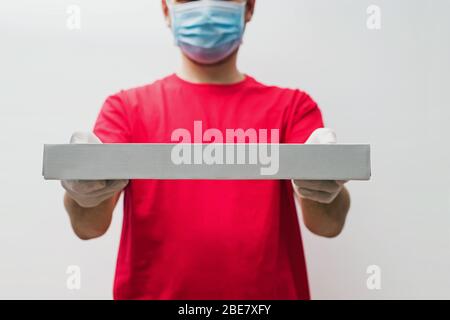 Der Lieferer trägt ein rotes T-Shirt, Schutzhandschuhe und eine Maske, die den Pizzakasten in der Nähe hält. Stockfoto