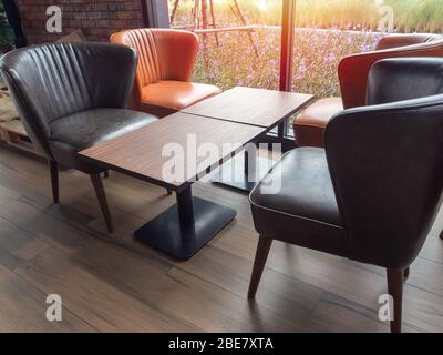 Lederstühle im Retro-Stil, orange und schwarze Farben und leerer Holztisch auf Holzboden im Café und Blumengarten Hintergrund draußen mit Sonnenschein. Stockfoto