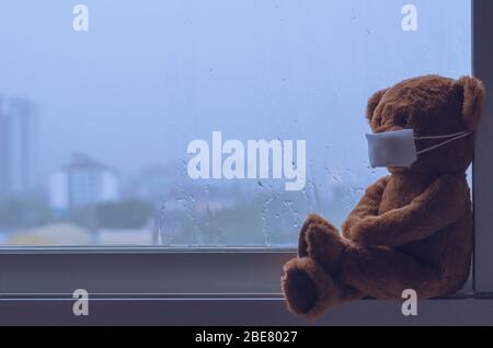 Teddybär trägt Maske am Fenster sitzen, während es in der Monsunzeit regnet. Bleiben Sie fern vom Virenkonzept zu Hause. Stockfoto