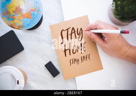 Hand halten Stift schreiben zu Hause bleiben Sie sicher auf braunem Papier mit blauen Globus auf Marmortisch bei home.protect Risiko, dass Coronavirus (covi Stockfoto