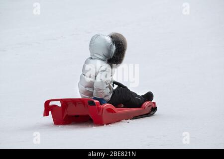 Ein junges Kind fährt mit einem roten Plastikschlitten auf einer Winterrutsche Stockfoto