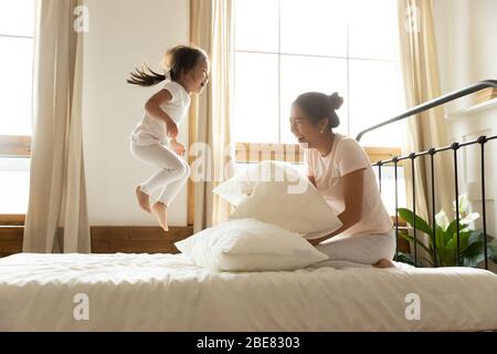 Sorglos kleine Tochter springen auf Bett, während vietnamesische Mutter lachen Stockfoto