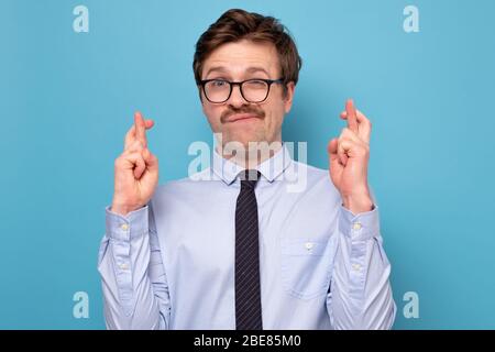 Junge kaukasische Geschäftsmann in lustigen Gläsern machen einen Wunsch isoliert auf blauem Hintergrund. Es ist meine letzte Chance. Stockfoto