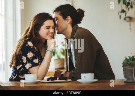 Schönes liebevolles Paar, das in einem Café sitzt und sich in einem Gespräch amüsst. Der Mann flüstert seiner Freundin an einem Kaffeetisch liebevoll ins Ohr. Stockfoto