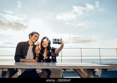 Frau, die Selfie mit Smartphone macht, mit ihrem Freund, der ein Eis im Freien hält. Paar, die Selfie an ihrem Datum. Stockfoto