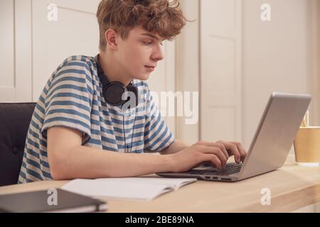 Ein netter Teenager in Kopfhörer benutzt einen Laptop und lernt an seinem Schreibtisch in seinem Zimmer Fernstudium wegen des epidemischen Coronavirus Stockfoto