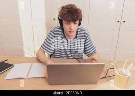 Ein netter Teenager mit Kopfhörern benutzt den Laptop und lernt am Schreibtisch in seinem Zimmer. Er macht Notizen mit einem Bleistift. Stockfoto