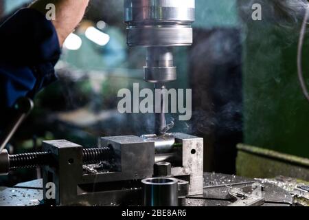 Industrieller Stahlbohrprozess mit Rauchnahaufnahme mit selektivem Fokus. Manuelle Zuführung von Hand. Werkstück im Schraubstock auf Tisch. Hoher Kontrast mit Hintergrundfarbe Stockfoto