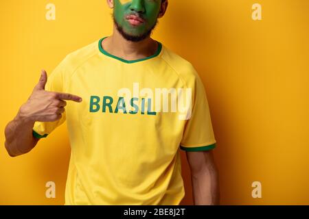 3/4-Ansicht des Fußballfans mit bemaltem Gesicht, das auf das T-Shirt zeigt, mit Brasilien-Schild auf Gelb Stockfoto