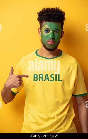 Seriöser afroamerikanischer Fußballfan mit bemaltem Gesicht, das auf T-Shirt mit brasilien-Schild auf Gelb zeigt Stockfoto