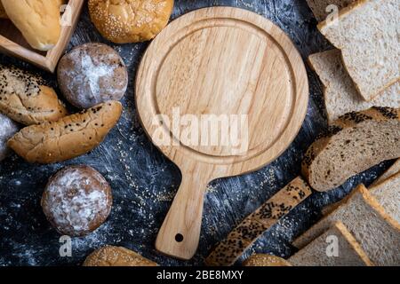 Ein leeres Holzbrett, das in der Mitte des Tisches von verschiedenen Brotsorten umgeben ist. Stockfoto