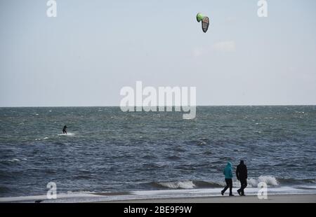 Trassenheide, Deutschland. April 2020. Zwei Leute laufen am Ostseestrand auf der Insel Usedom entlang, mit einem Kitesurfer im Hintergrund. Um die Ausbreitung des Corona-Virus zu verlangsamen, hat die Bundesregierung das öffentliche Leben erheblich eingeschränkt. Quelle: Stefan Sauer/dpa/Alamy Live News Stockfoto