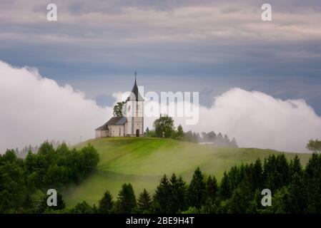 Jamnik Kirche auf einem Hügel im Frühling, neblig Wetter bei Sonnenuntergang in Slowenien, Europa. Berglandschaft kurz nach dem Frühlingsregen. Slowenische Alpen. Stockfoto