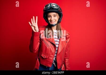 Junge schöne Brünette Motorradfahrerin Frau trägt Motorradhelm und rote Jacke zeigt und zeigt mit den Fingern Nummer vier beim Lächeln con Stockfoto