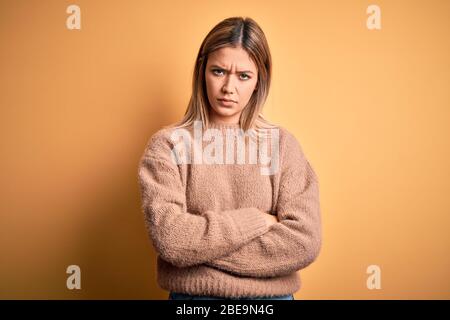 Junge schöne blonde Frau trägt braunen Pullover über gelb isoliert Hintergrund skeptisch und nervös, missbilligend Ausdruck im Gesicht mit gekreuzten ein Stockfoto