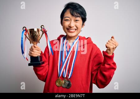 Junge schöne asiatische Mädchen Gewinner mit Trophäe trägt Medaillen auf weißem Hintergrund schreien stolz und feiern Sieg und Erfolg sehr aufgeregt Stockfoto