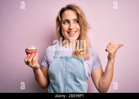 Junge schöne blonde Frau eatimg Schokolade Cupcake über isoliert rosa Hintergrund zeigen und mit dem Daumen bis zur Seite mit glücklichem Gesicht smi zeigen Stockfoto