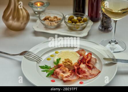 Warmer Ziegenkäse mit Honig und Speck auf einem weißen Gericht serviert. Rosetten mit Mandeln und grünen Oliven und ein Glas Weißwein auf dem Tisch. Stockfoto