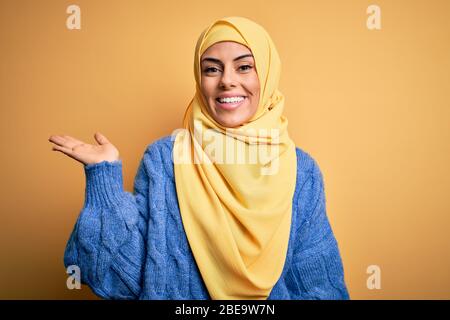 Junge schöne Brünette muslimische Frau trägt arabischen Hijab über isolierten gelben Hintergrund lächelnd fröhlich präsentieren und zeigen mit Handfläche lo Stockfoto