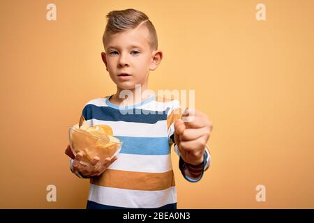 Junge kleine kaukasische Kind essen ungehitzige Kartoffeln Chips Chips über gelbem Hintergrund verärgert und frustriert schreien vor Wut, verrückt und schreien Stockfoto